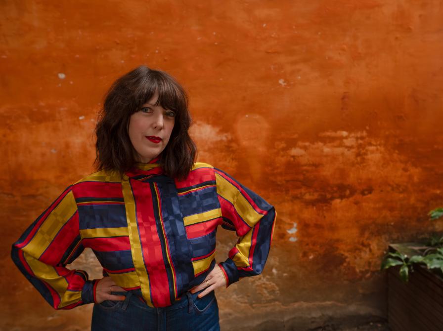 Kvinde der ser ind i kameraet, foran en orange murstens mur, iført en farvesteålende jakke.